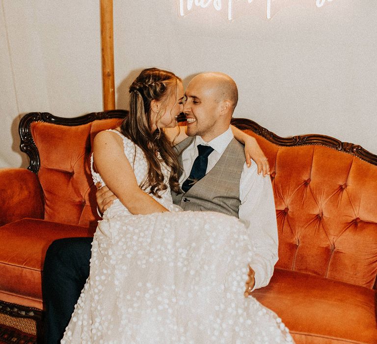 The bride sits on the lap of the groom on a red velvet sofa with a custom made white neon light wedding sign in the marquee 