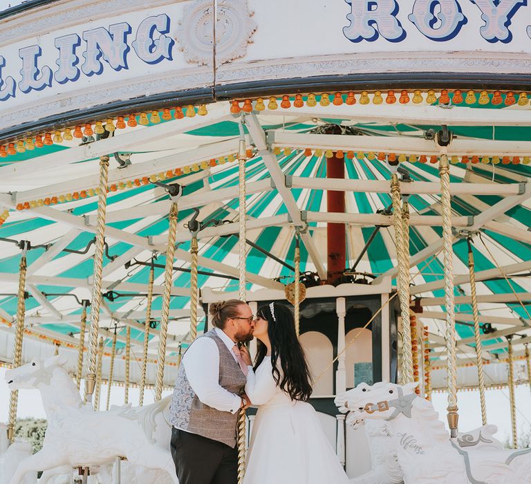 The bride in a long sleeve princess wedding dress with the groom in a light grey checkered waistcoat sharing a kiss on the carousel 