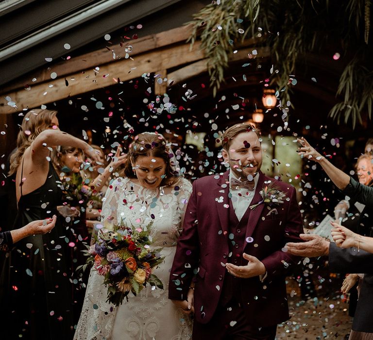 The groom in a three piece burgundy suit with dried flower buttonhole has confetti moment with the bride in a boho lace wedding dress 