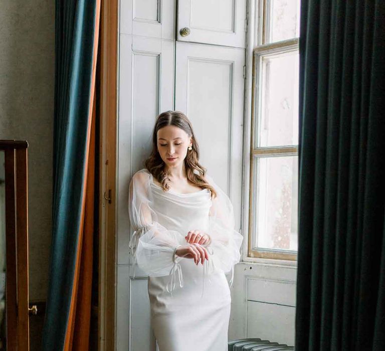 Bride stading in St Giles House wearing long sheer puff sleeve satin wedding dress with cowl neck 