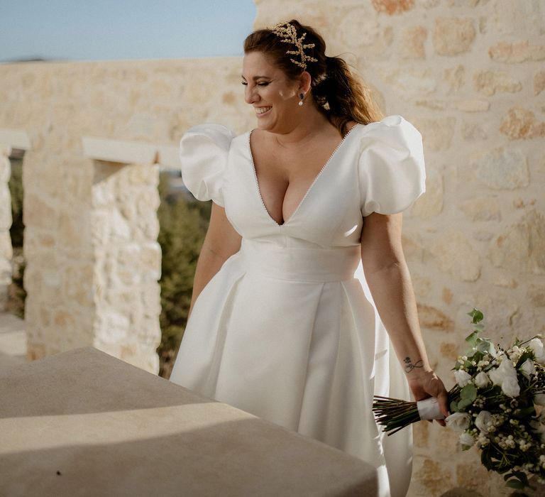 Bride laughing and holding bridal bouqet that features wihte berries, italian ranunculus and eucalyptus whilst wearing a stain puff-sleeved wedding dress, peal earrings and gold leaf bridal grown