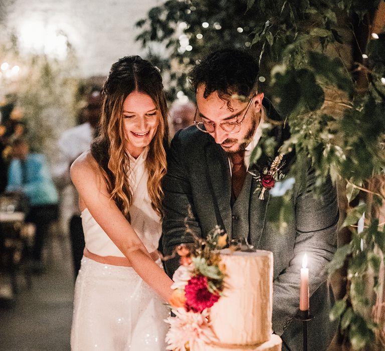 Groom in three piece suit cutting the pale pink wedding cake with the bride in a sparkly wedding reception outfit for November wedding 