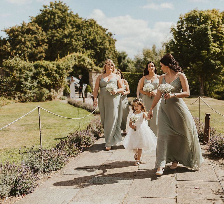 Bridesmaids in matching sage green bridesmaid dresses walk to the venue with the flower girl in a white dress 