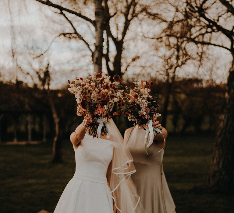 The bride and maid of honour hold their dried flower bouquets tied with white ribbon over their faces 