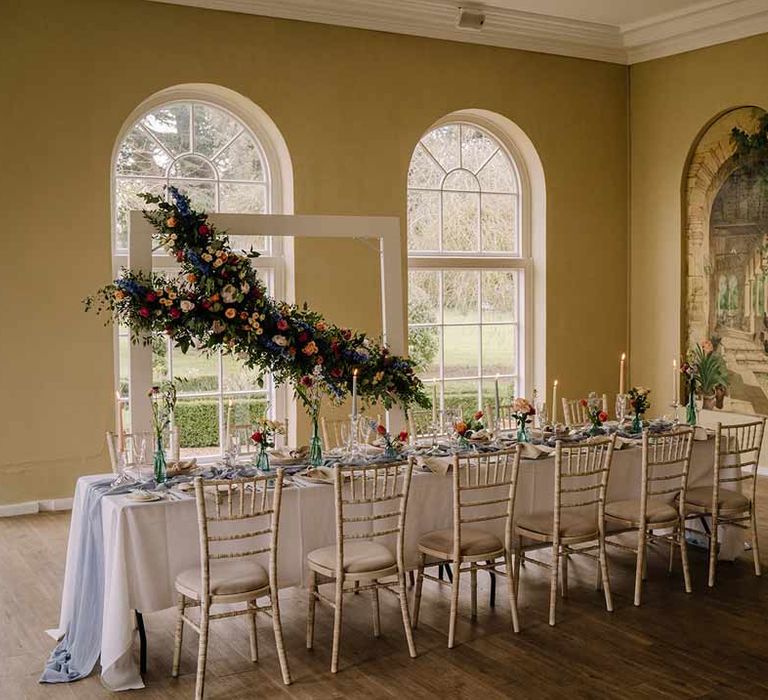 Classic wedding tablescape with white wedding tablecloth, cornflower blue wedding table runner, bright blue glass bottles with dried flowers, pastel tapered candles and spring floral arrangements with suspended spring floral decor 