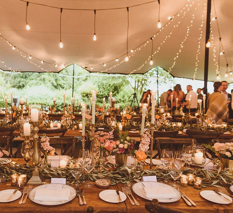 Marquee wedding with fairy lights and festoon lighting for outdoor wedding breakfast with pastel taper candles and bright table funner made of foliage and flowers 