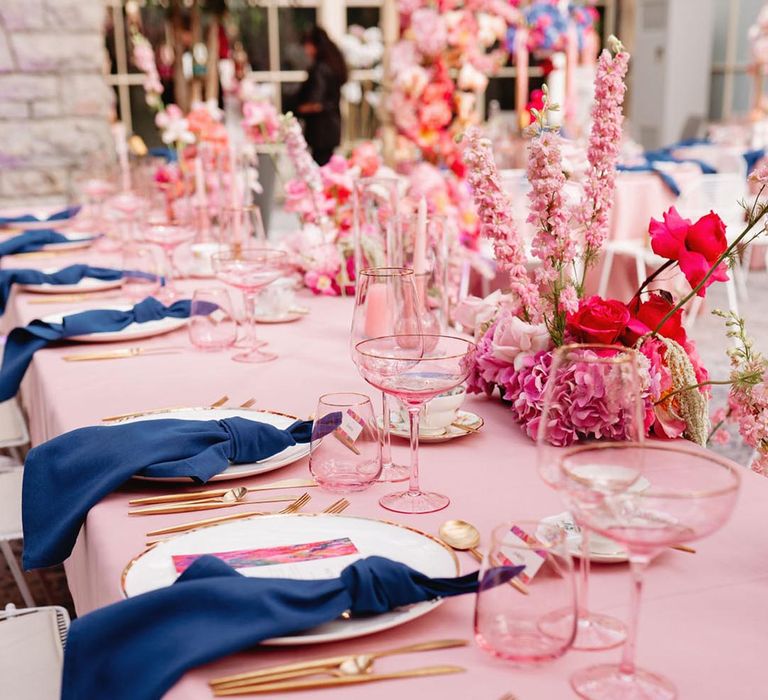 White plate with gold edges and gold cutlery with dark blue napkin and pink tablecloths and flowers 