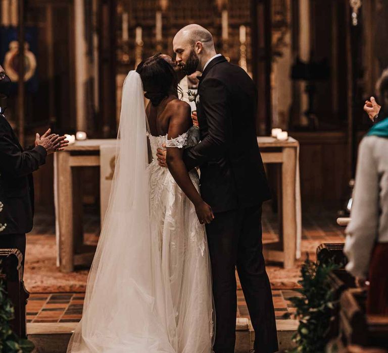 Bride in sweetheart neckline lace wedding dress with detachable off the shoulder sleeves standing with groom in classic black three piece grooms suit with bow tie and white rose and foliage boutonniere in church 