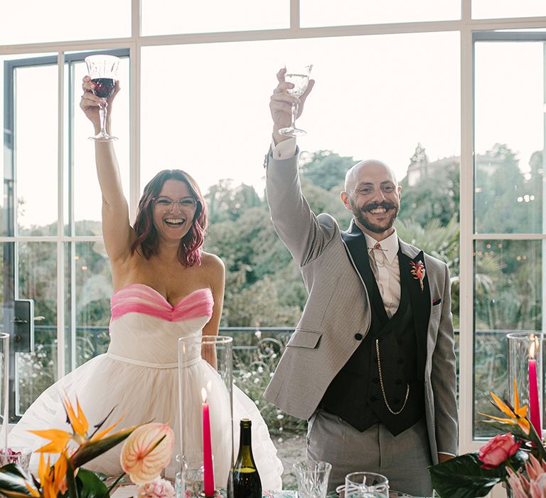 bride wearing glasses in a strapless wedding dress and groom in a grey suit raising a toast at their italian villa wedding