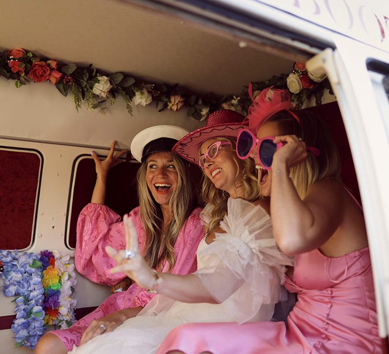 Bride and bridal party sit in Vintage Camper Booth photo booth with pink props for pictures 