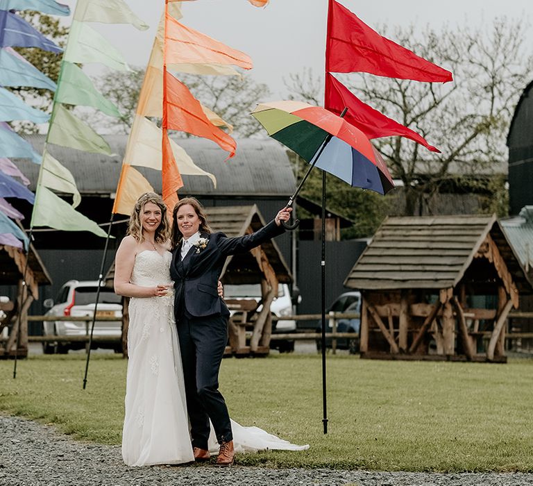 bride in a lace wedding dress and bride in a navy suit stand in front of colourful festival flags at Stanford Farm wedding 