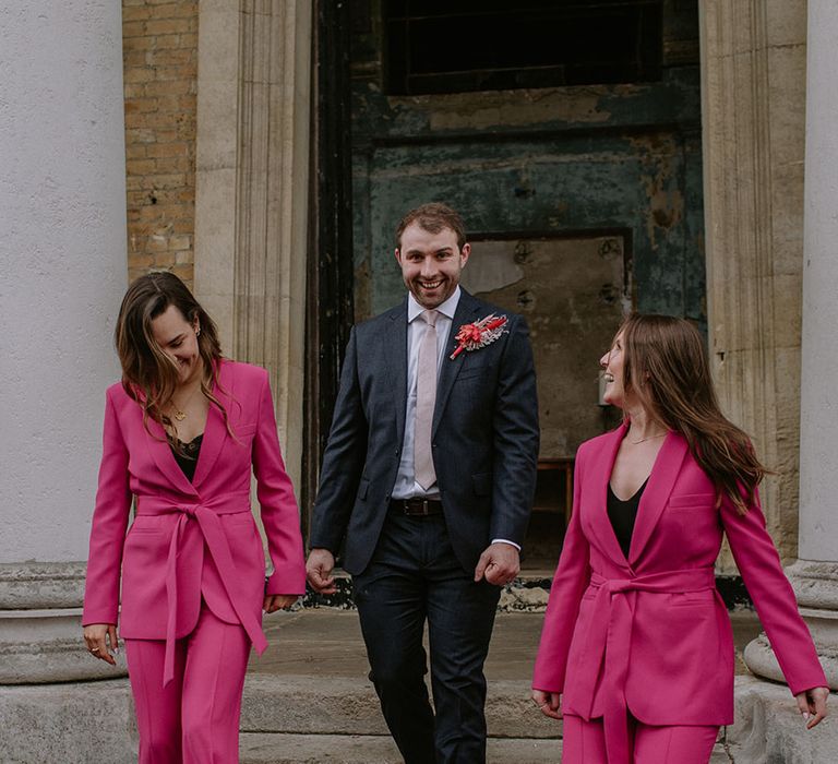 Groom in navy suit with groomswomen in hot pink matching suits as they walk down the stairs at the London wedding venue 