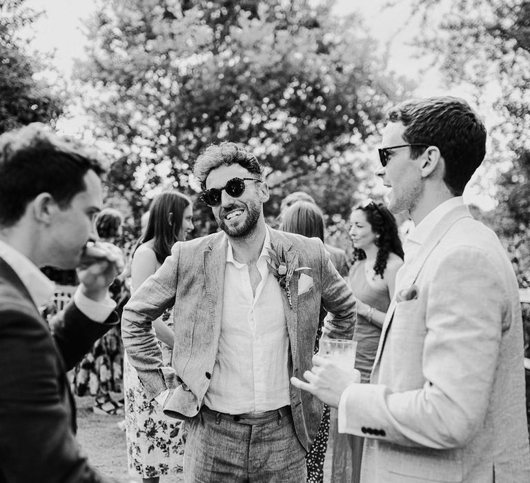 The groom in a grey suit socialises and shares drinks with guests at wedding 