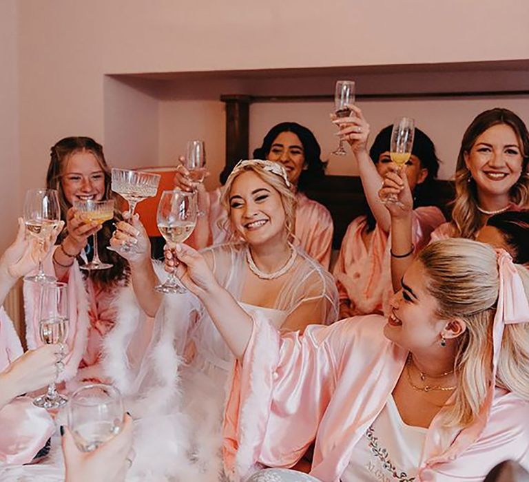 Bridal party sitting on a bed for group photo while cheersing prosecco in pink satin robes by Marina Walker Photography 