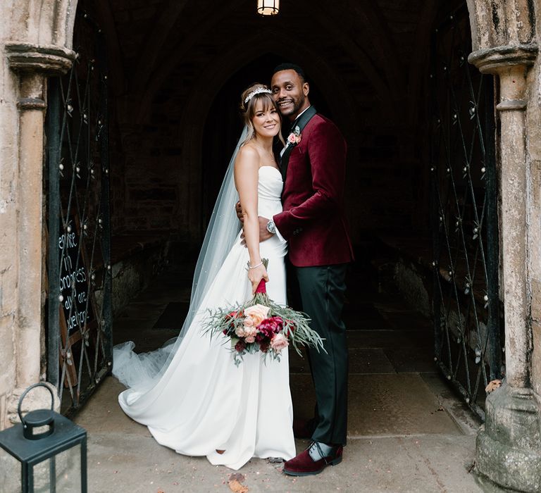 The groom in a red velvet suit jacket embraces the bride in a strapless gown at their luxury castle wedding venue 