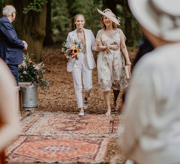 Bride in white wedding suit and lace bodysuit walks down the aisle with the mother of the bride 