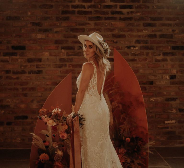 Bride in lace wedding dress standing in front of an orange board and floral altar