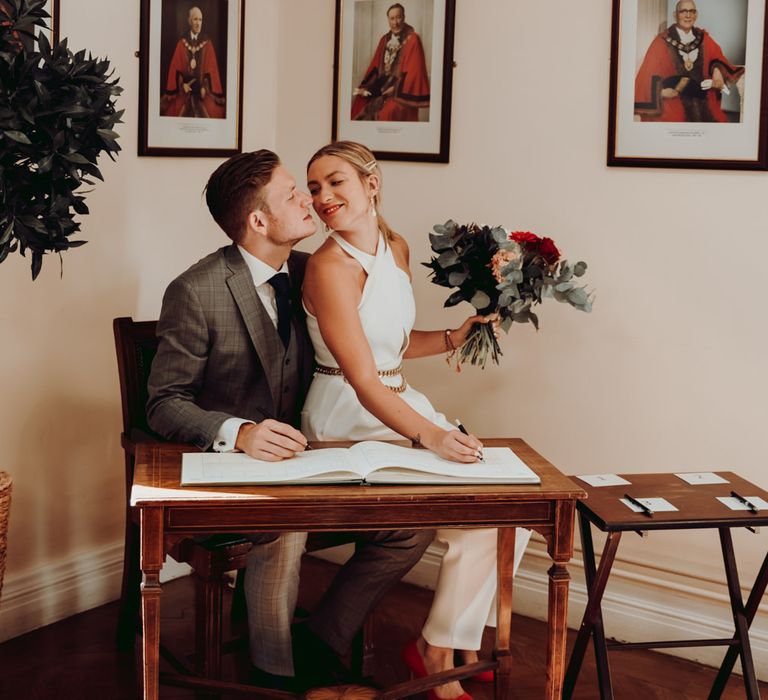 Bride in Massimo Dutti jumpsuit sitting on her grooms lap signing the register 