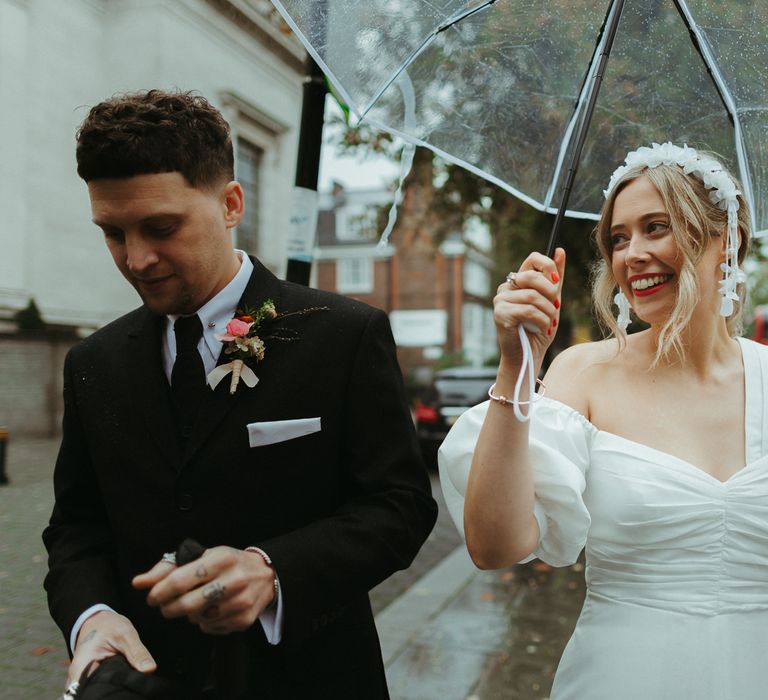 Bride and groom walk in London on a rainy day with a pink boutonnière and ruched wedding dress