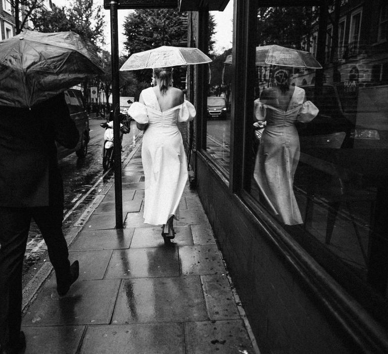 Bride walks with clear umbrella in a puff sleeve ruched wedding dress alongside groom with black umbrella and suit