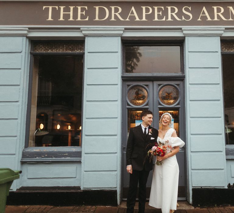 Smiling bride and groom pose in front of pub in London in puff sleeve wedding dress and bright flowers 