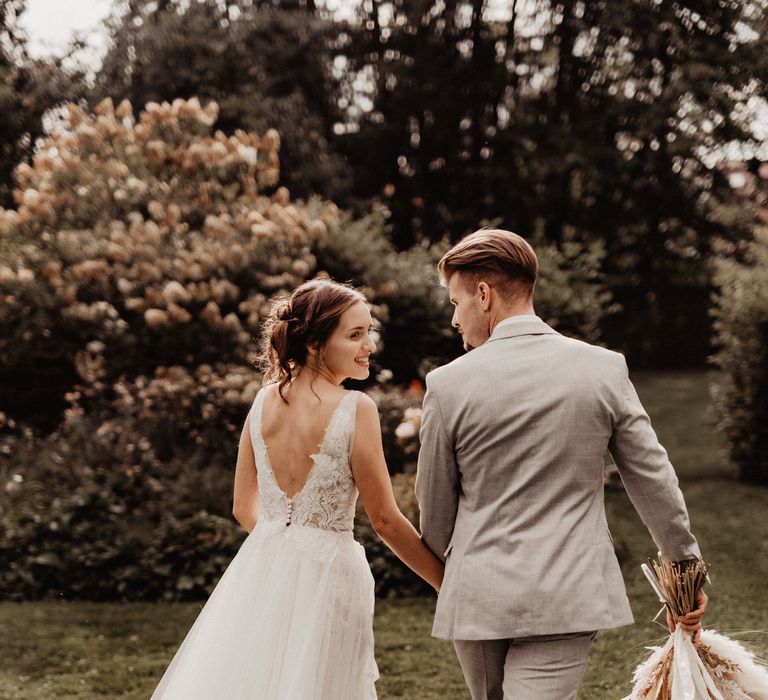 Bride in backless Watters wedding dress walking in garden with Groom