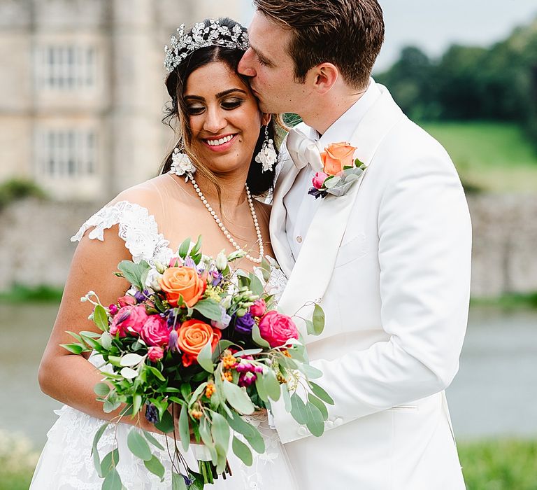 Groom in a Tom Ford White Tuxedo jacket kissing his bride on the head in a princess wedding dress and bridal crown whilst holding a colourful bouquet 