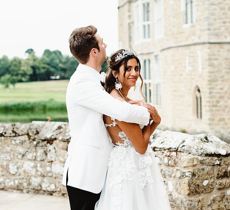 Groom in a white tuxedo jacket embracing his princess bride in an off the shoulder wedding dress outside their castle wedding venue 