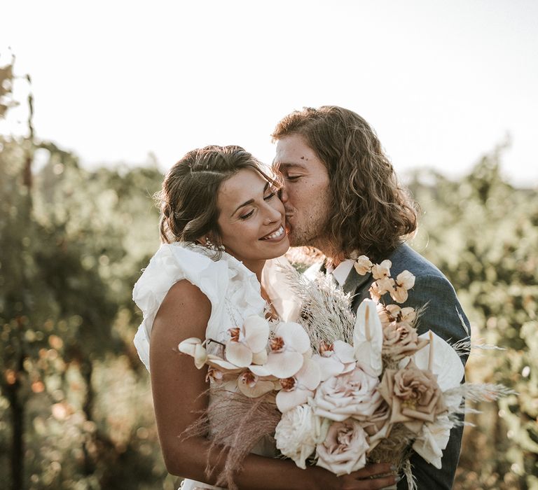 Groom with shoulder length curly hair in a navy blue suit kissing his brides cheek in a ruffle sleeve wedding dress holding an orchid wedding bouquet 