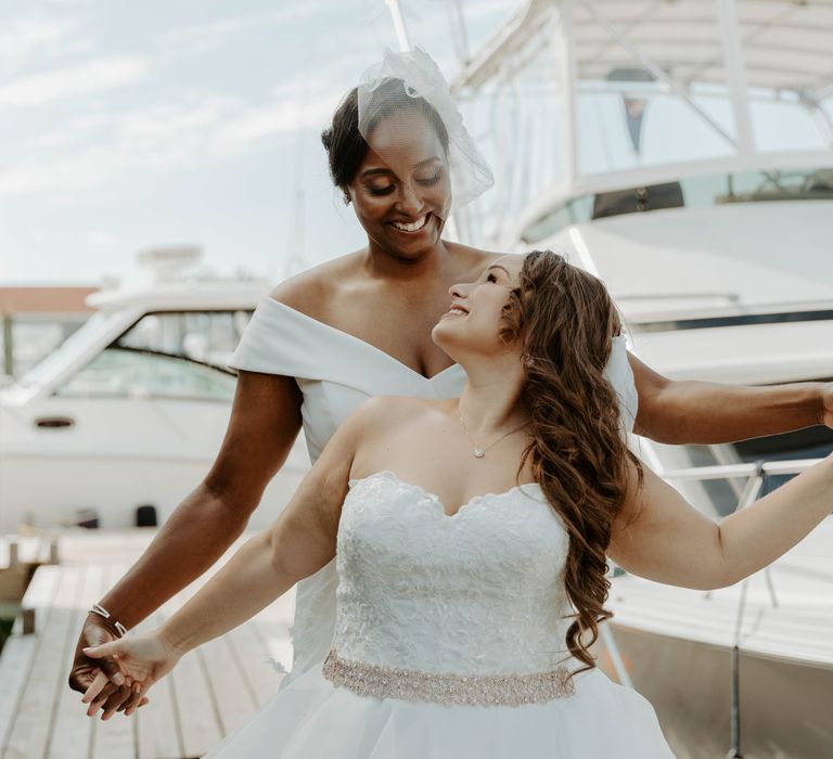 Black bride in Bardot wedding jumpsuit look at her bride in a strapless wedding dress whilst they hold hands together 