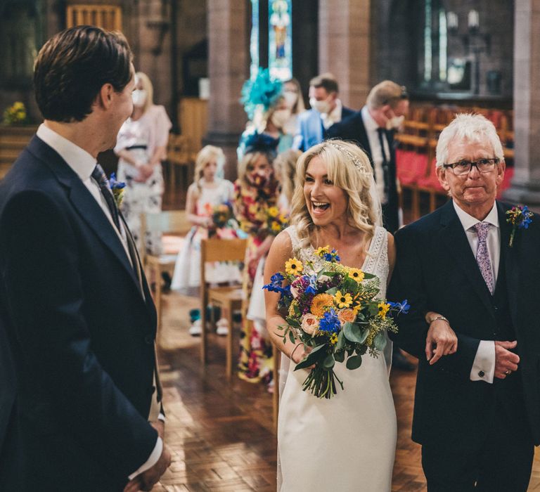 Bride holding colourful bouquet smiles at groom whilst being walked up the aisle by her father in church ceremony