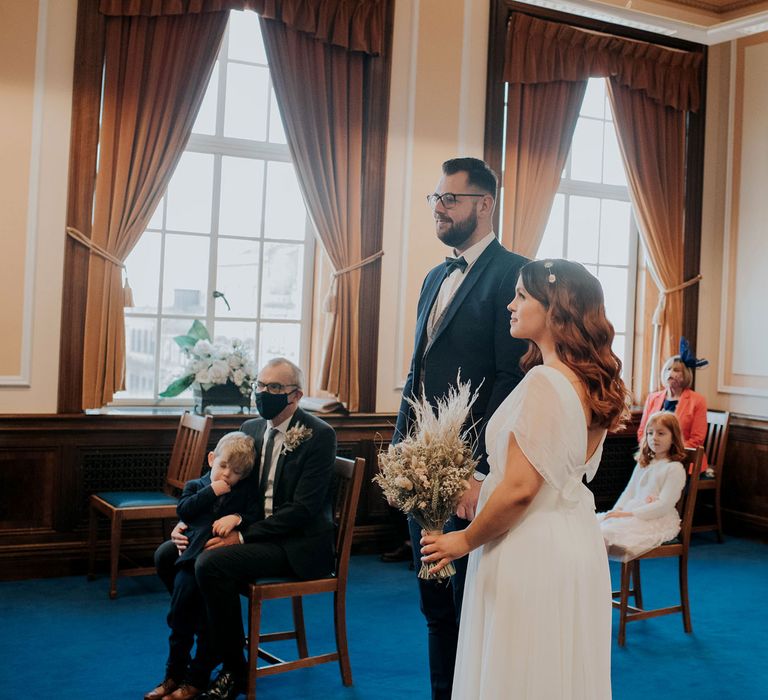 Bride in white dress and celestial headband holding dried flower bouquet during civil town hall ceremony standing next to groom in navy suit