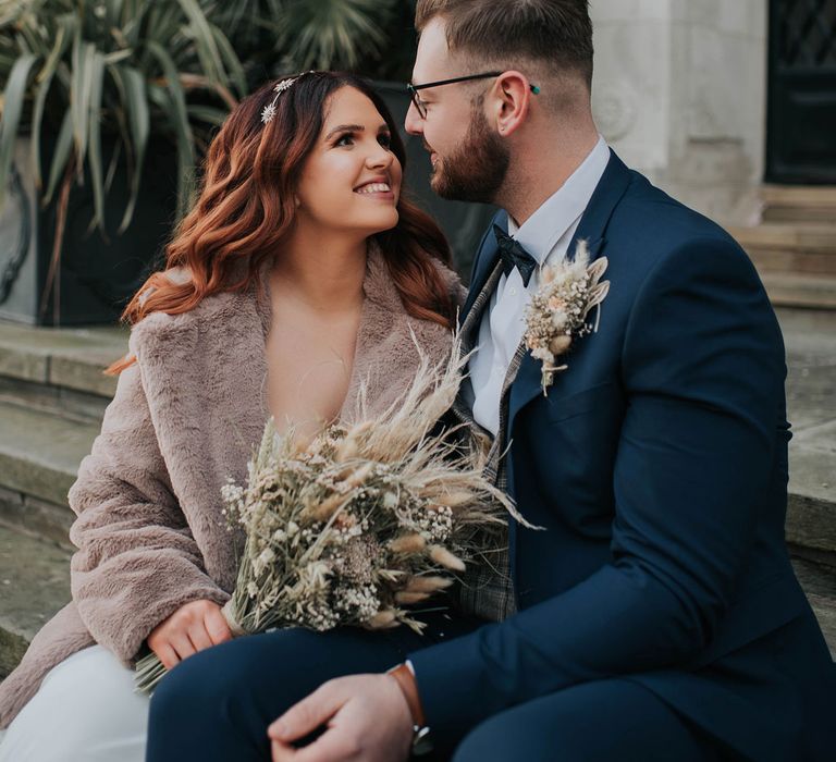 Bride in fur coat and celestial headband holding dried flower bouquet smiles at groom in navy suit whilst sat on steps