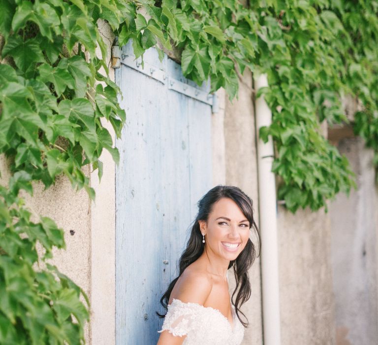 Dark haired bride with loose curls and wearing off the shoulder wedding gown leans against blue wooden door 