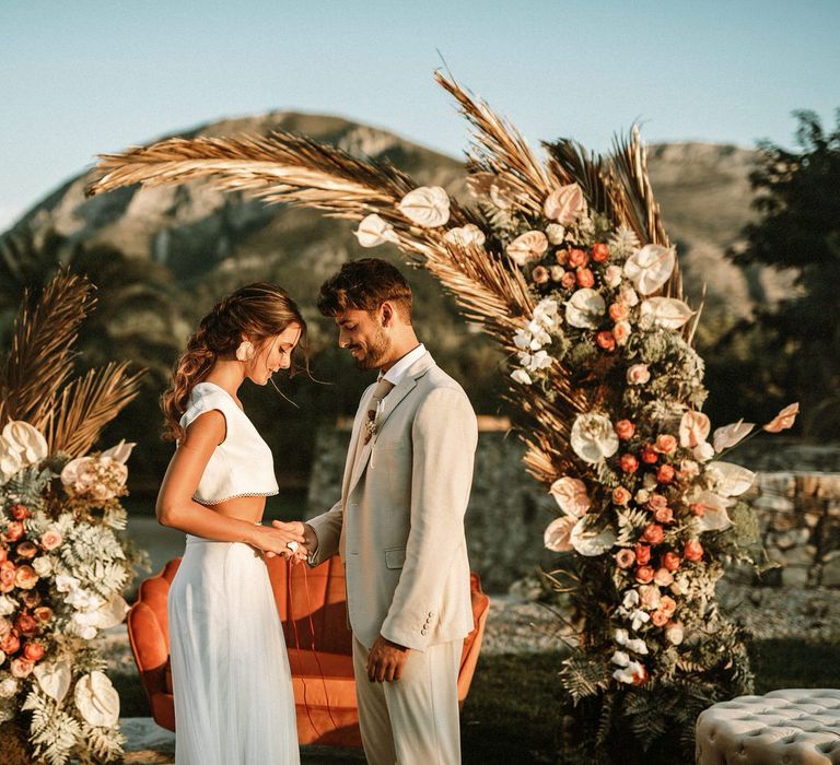 Outdoor hand fasting ceremony for boho bride and groom at their floral moon gate and wicker rug altar 