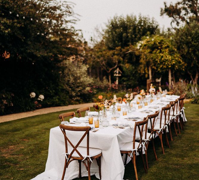 Intimate garden party wedding reception with wooden table and chairs, white table cloth and yellow glassware. 