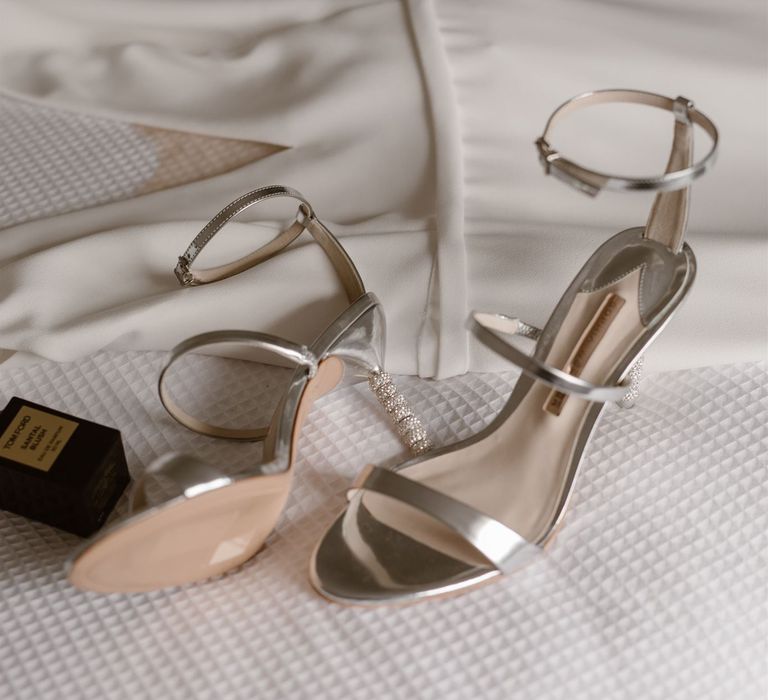 Silver Sophia Webster heeled sandals with three straps and studded heel on white bedspread with black bottle of Tom Ford Eau de Parfum