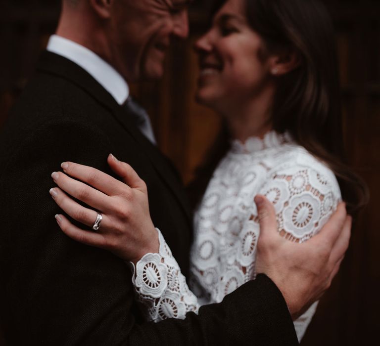 Smiling groom in brown woollen blazer and smiling bride in long sleeved lace top Self Portrait wedding dress and silver wedding ring hold each others arms