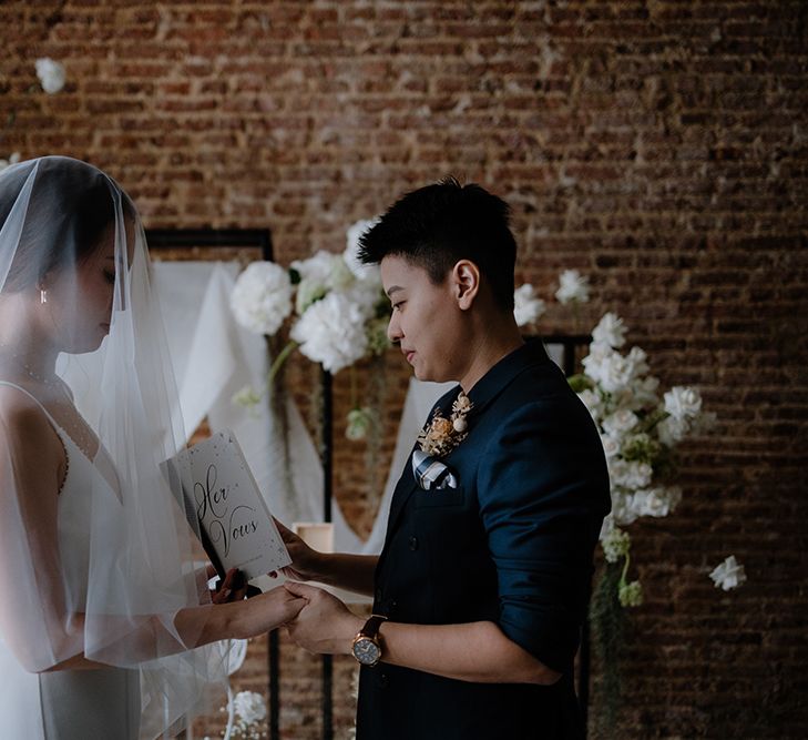 Asian bride in a navy suit jacket reading her vows from a vow booklet 