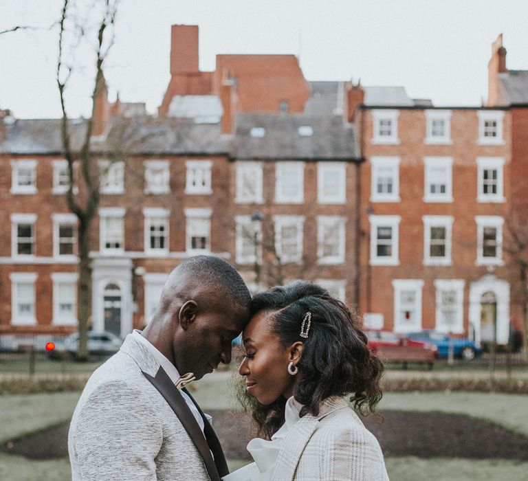 Groom in grey suit jacket with black lapels and bow tie stands with his head touching bride with curled hair and grey checked jacket