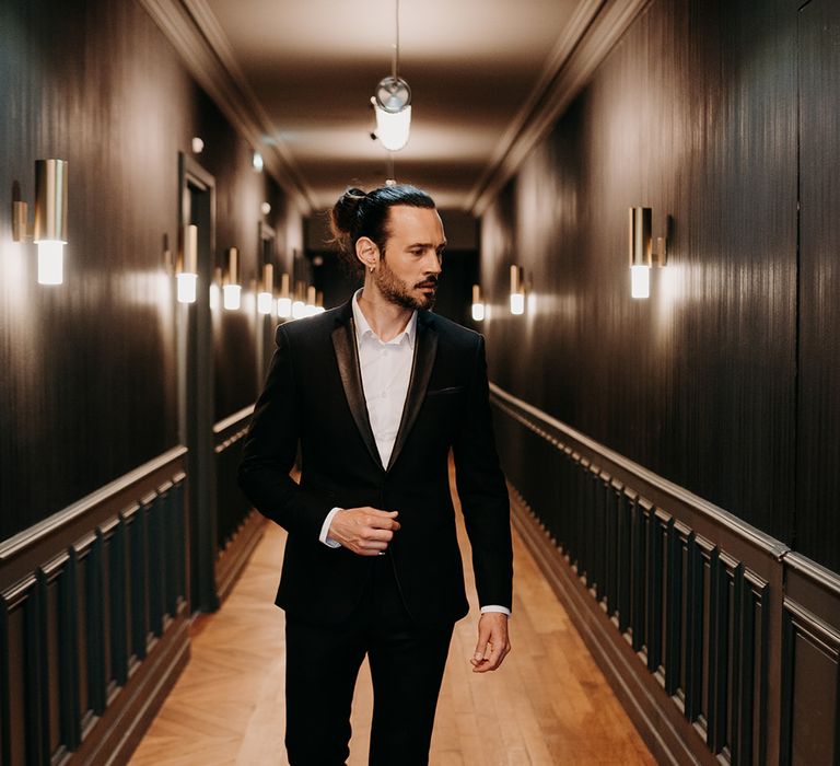 Groom walks through hallway wearing slim & tailored black suit with hair slicked back