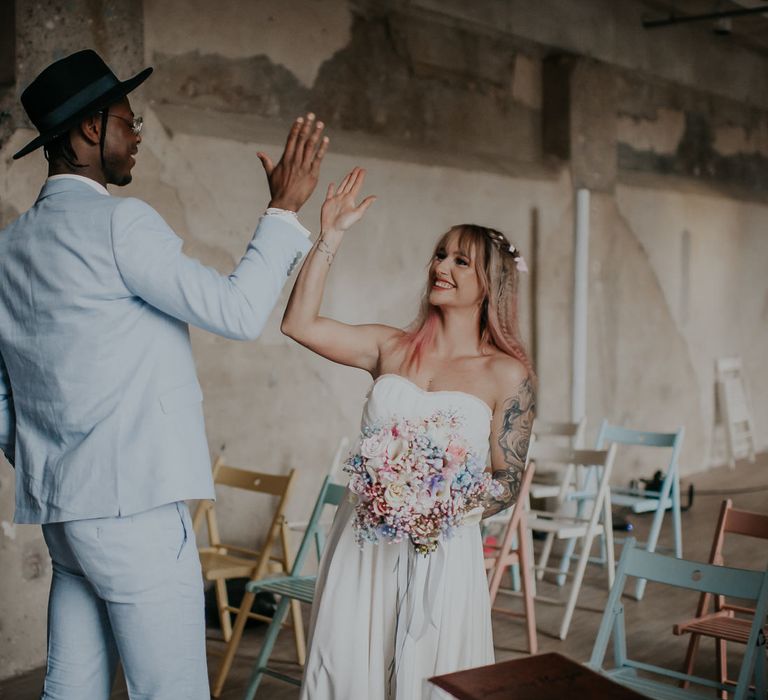 Bride with pink ombre hair and tattoos wearing a crop top and skirt giving her groom in a blue suit a high five at the altar 