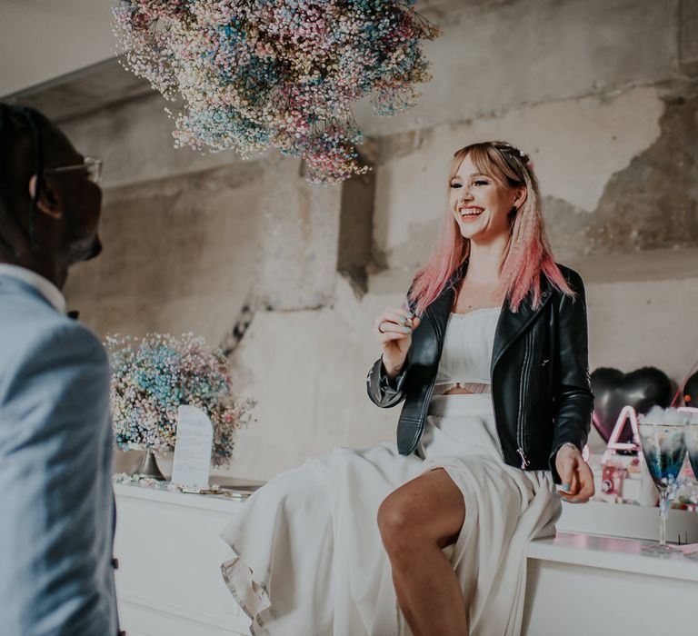 Bride with pink ombre hair wearing a leather jacket sitting on the bar under a pastel flower cloud 