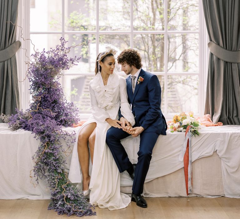 Bride & groom sit on table next to twisted lavender whilst holding hands and looking lovingly at one another