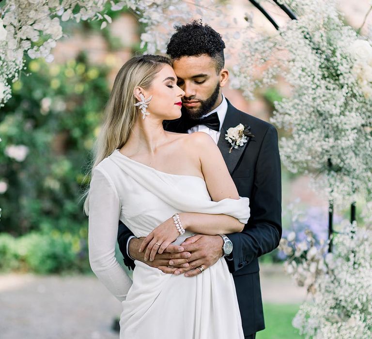 Groom in a black tuxedo embracing his bride in a one shoulder wedding dress with long sleeve and statement earring 