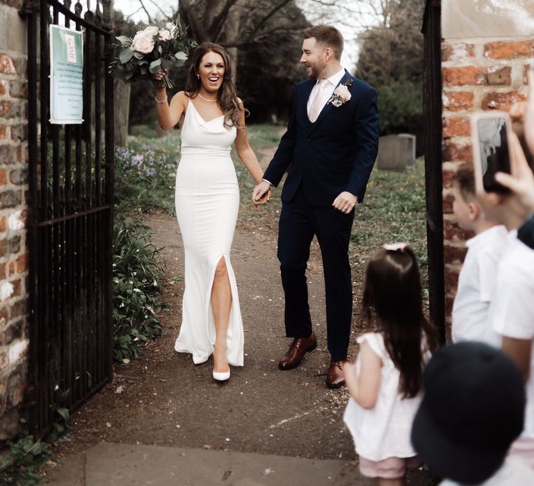 Bride and groom leaving their church wedding, the bride is wearing a white slip wedding dress and holding her bouquet in the air