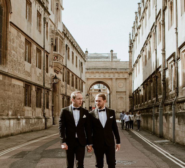 A gay couple walk down a street for wedding portraits. They wear black tuxedos. 