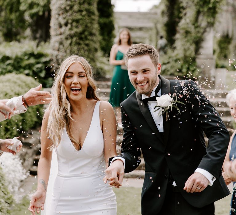 Bride & groom walk together holding hands whilst confetti is thrown around them in gardens