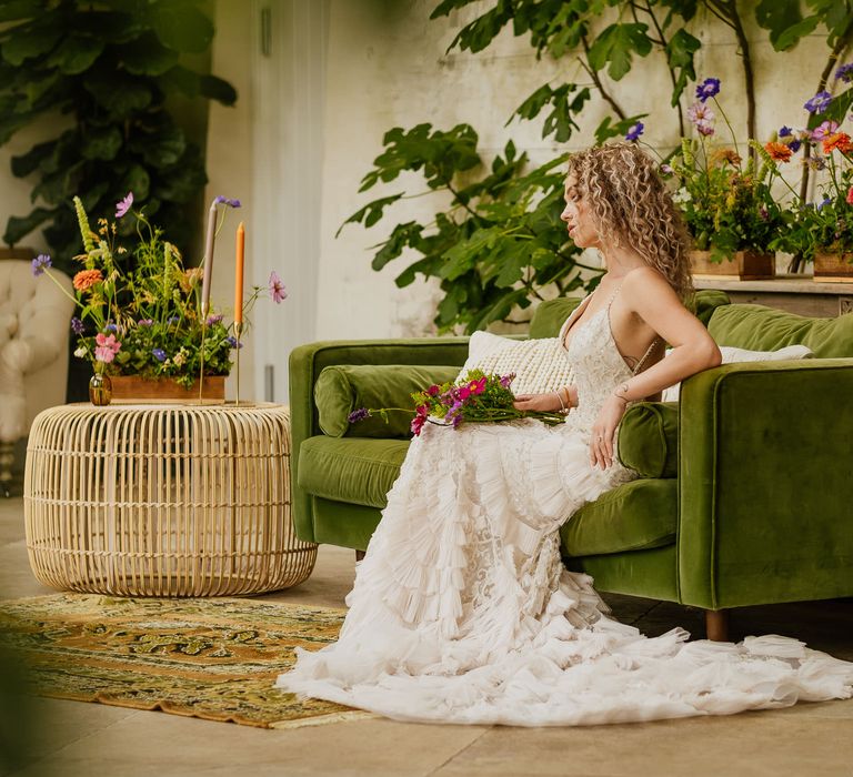 Blonde curly haired bride sitting on a green velvet sofa in a embellished wedding dress surrounded by colourful wildflower decor 