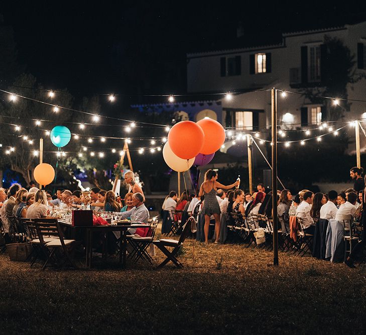 Wedding reception during the nighttime in Menorca with fairy lights strung above wooden tables and colourful balloons